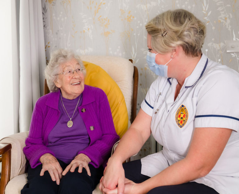 Older lady in a purple jumper sat in a chair with a yellow cushion is smiling to her physiotherapist whilst they discuss her problems.