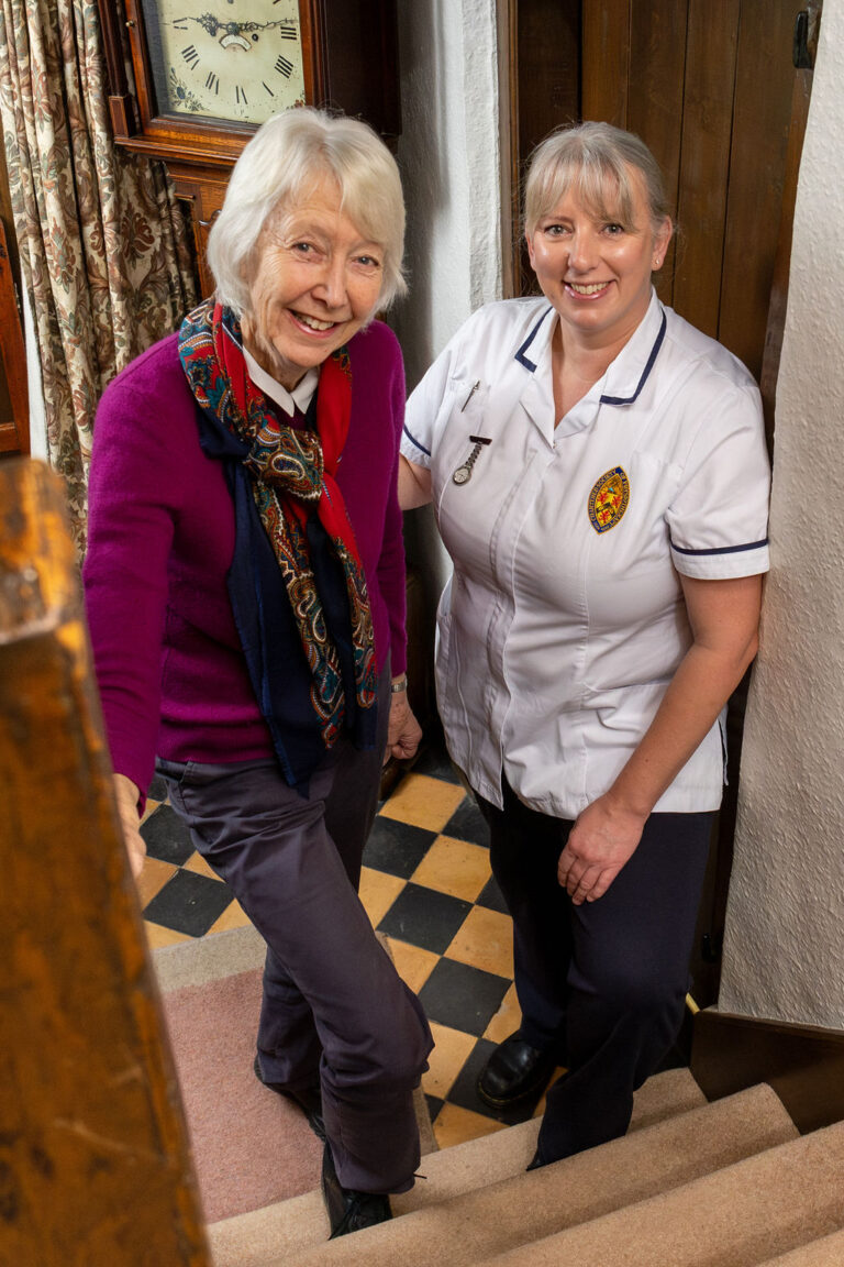 Older lady in a colourful scarf and purple jumper is standing at the foot of the stairs with a physiotherapist, both are smiling.