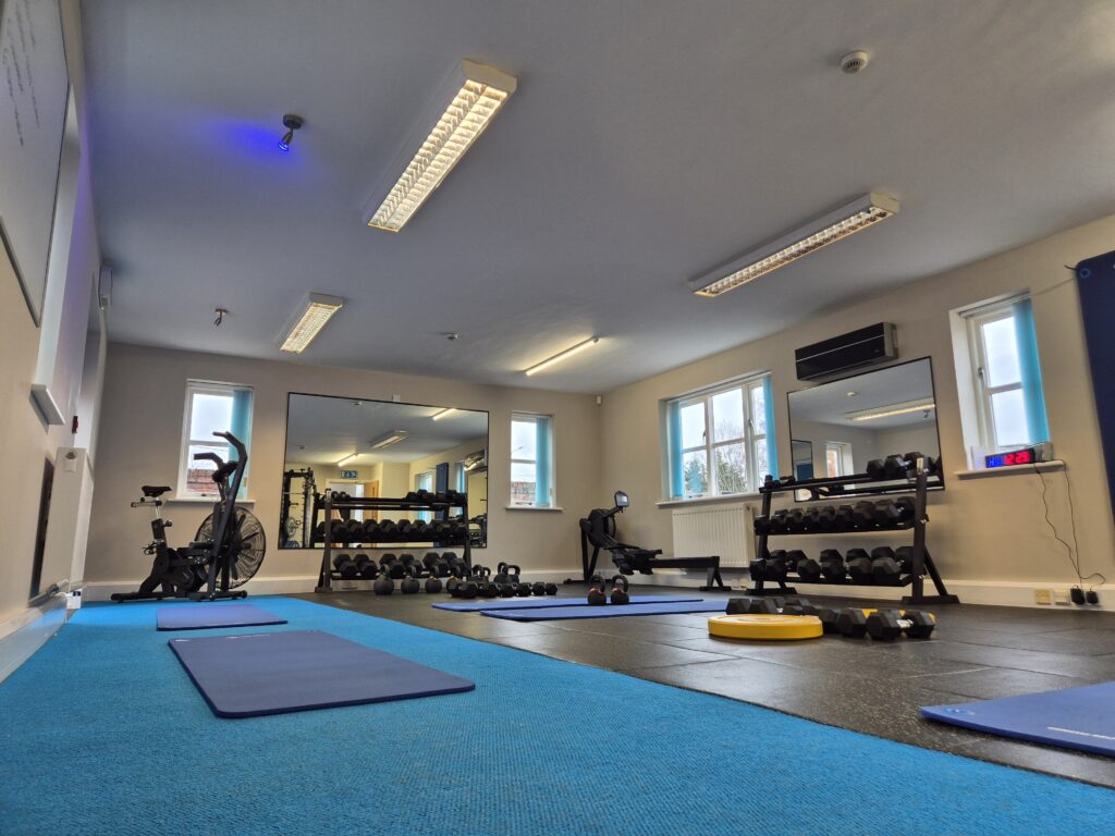 A wide-angle view of the Houlbrook Physiotherapy rehabilitation gym featuring blue exercise mats, kettlebells, and dumbbells on a black rubber floor, with a functional cable machine, rowing machine, and air bike in a bright, professional studio.