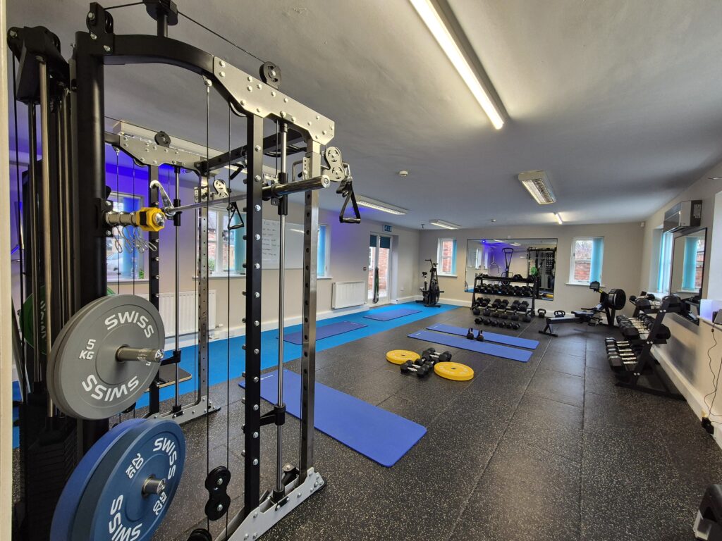 A modern, well-equipped rehabilitation gym featuring a multi-functional cable machine, free weights, dumbbells, and blue exercise mats on a professional rubber floor.
