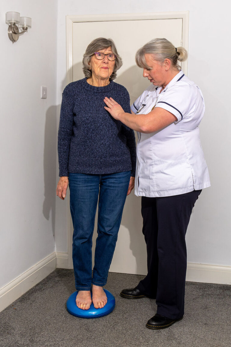 Lady in a blue jumper and jeans is balancing on a blue wobble cushion, her physiotherapist supporting her at the side.